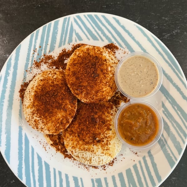Three idlis topped with red spice powder, served with two chutneys on a striped plate.