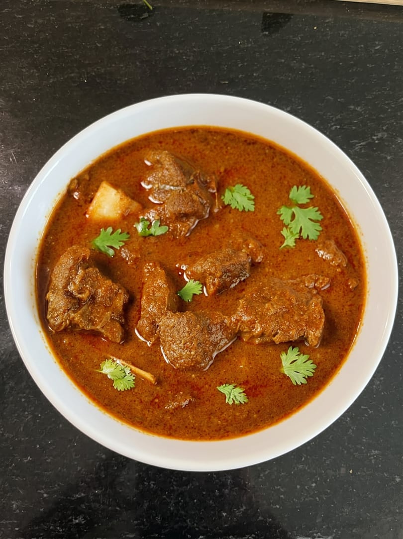 A white bowl filled with rich, reddish-brown curry, garnished with fresh green cilantro leaves.