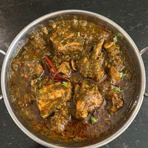 A close-up of a metal bowl filled with green chicken curry, garnished with red chili and cilantro.