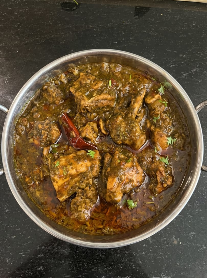 A close-up of a metal bowl filled with green chicken curry, garnished with red chili and cilantro.