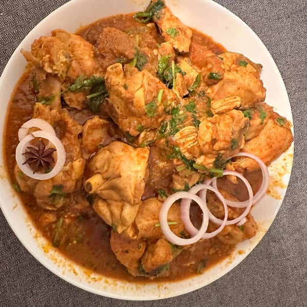 Chicken curry with cilantro, star anise, and red onion slices in a white bowl.